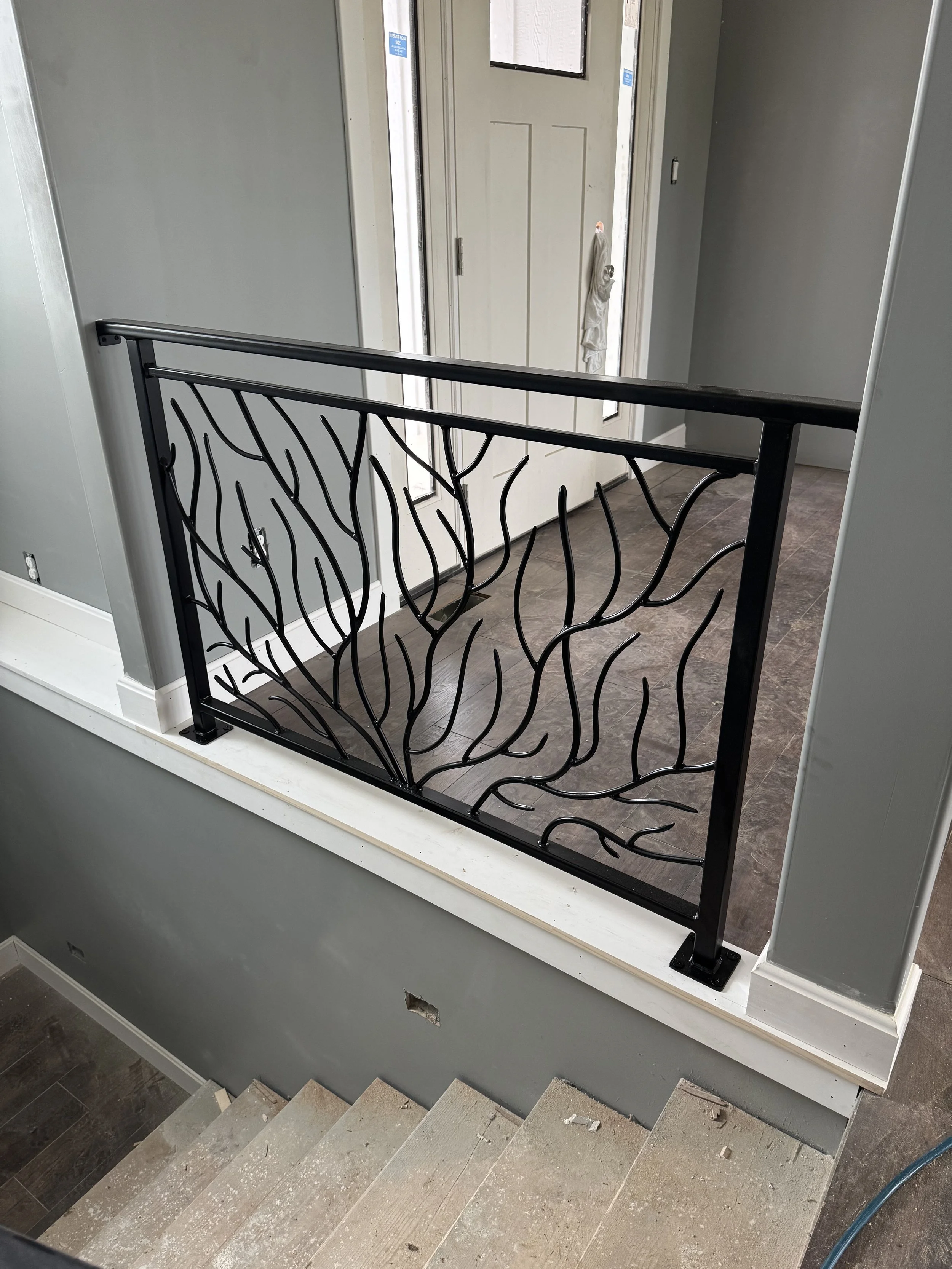 Interior view of an unfinished staircase and landing with a decorative black wrought iron railing, an open front door, and stairs leading downward with exposed edges and construction debris.