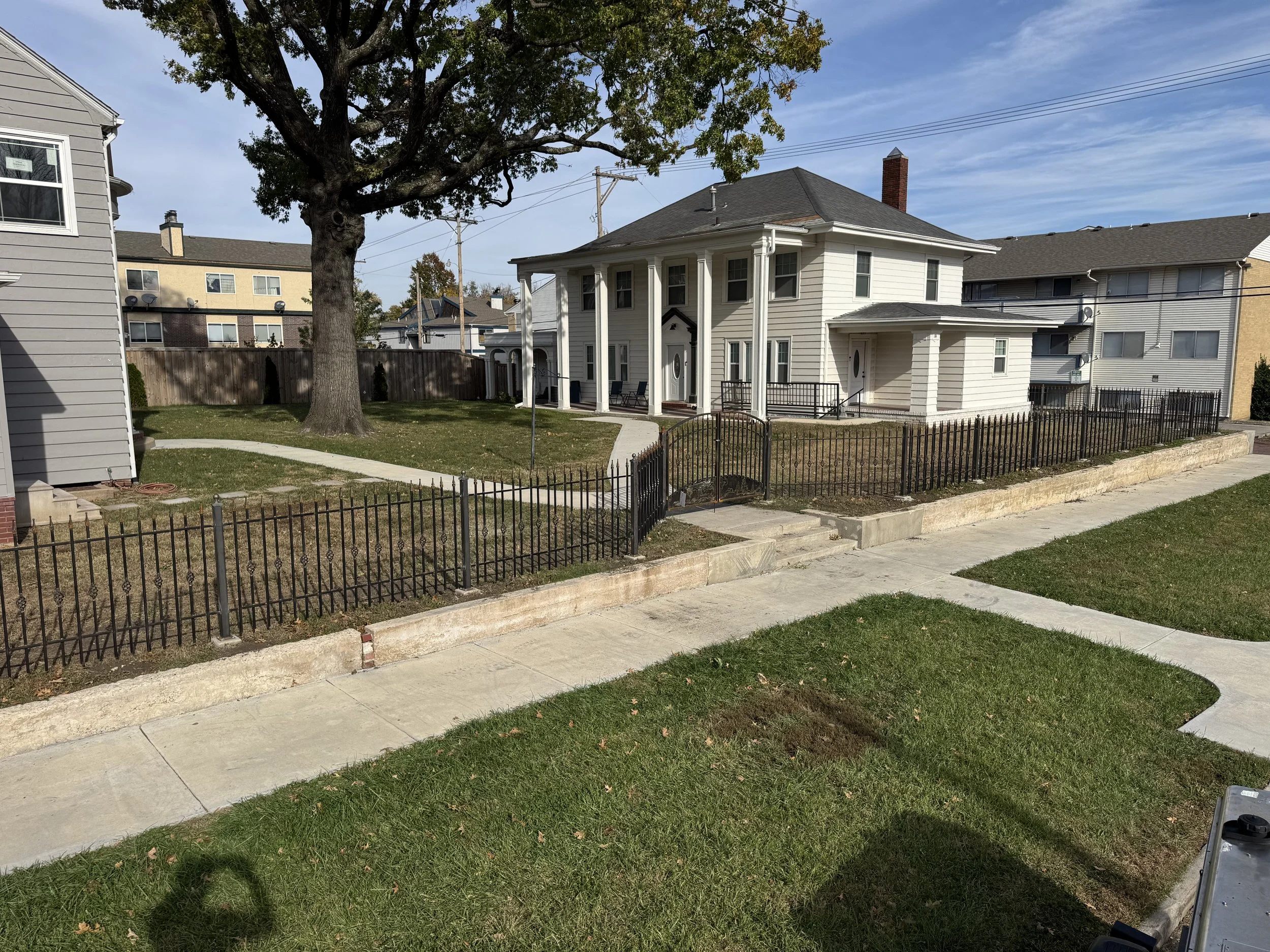 View of a residential area showing a white house with columns, a large tree, a small fenced yard, neighboring houses, a sidewalk, and utility poles under a partly cloudy sky.