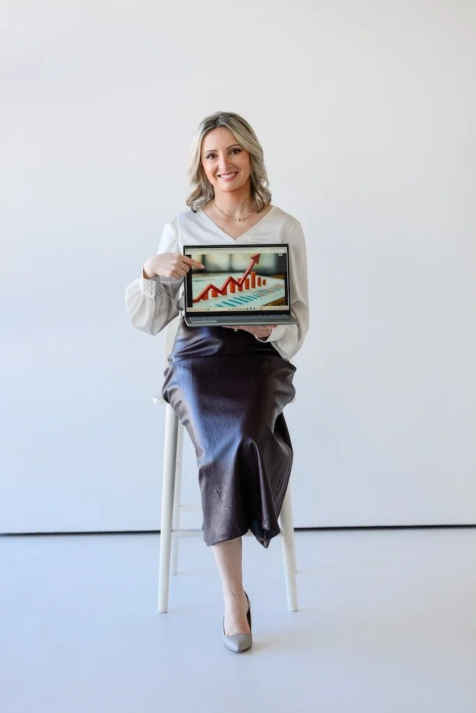 A woman sitting on a white stool, wearing a white blouse and leather skirt, holding a laptop displaying a rising bar graph with an upward arrow.