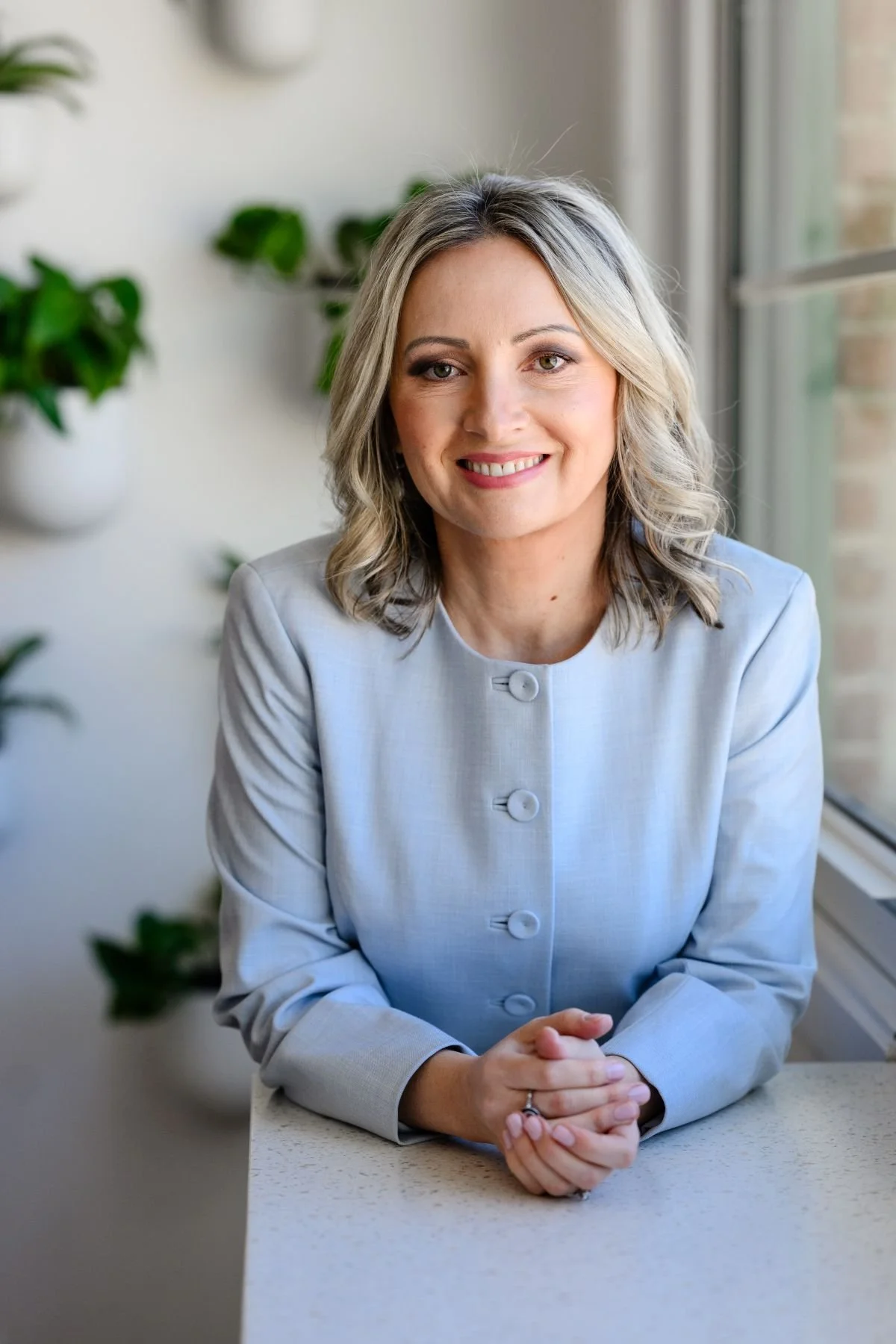 A woman with blonde hair and green eyes sitting at a countertop near a window, wearing a light blue blazer, smiling at the camera with her hands clasped in front of her.
