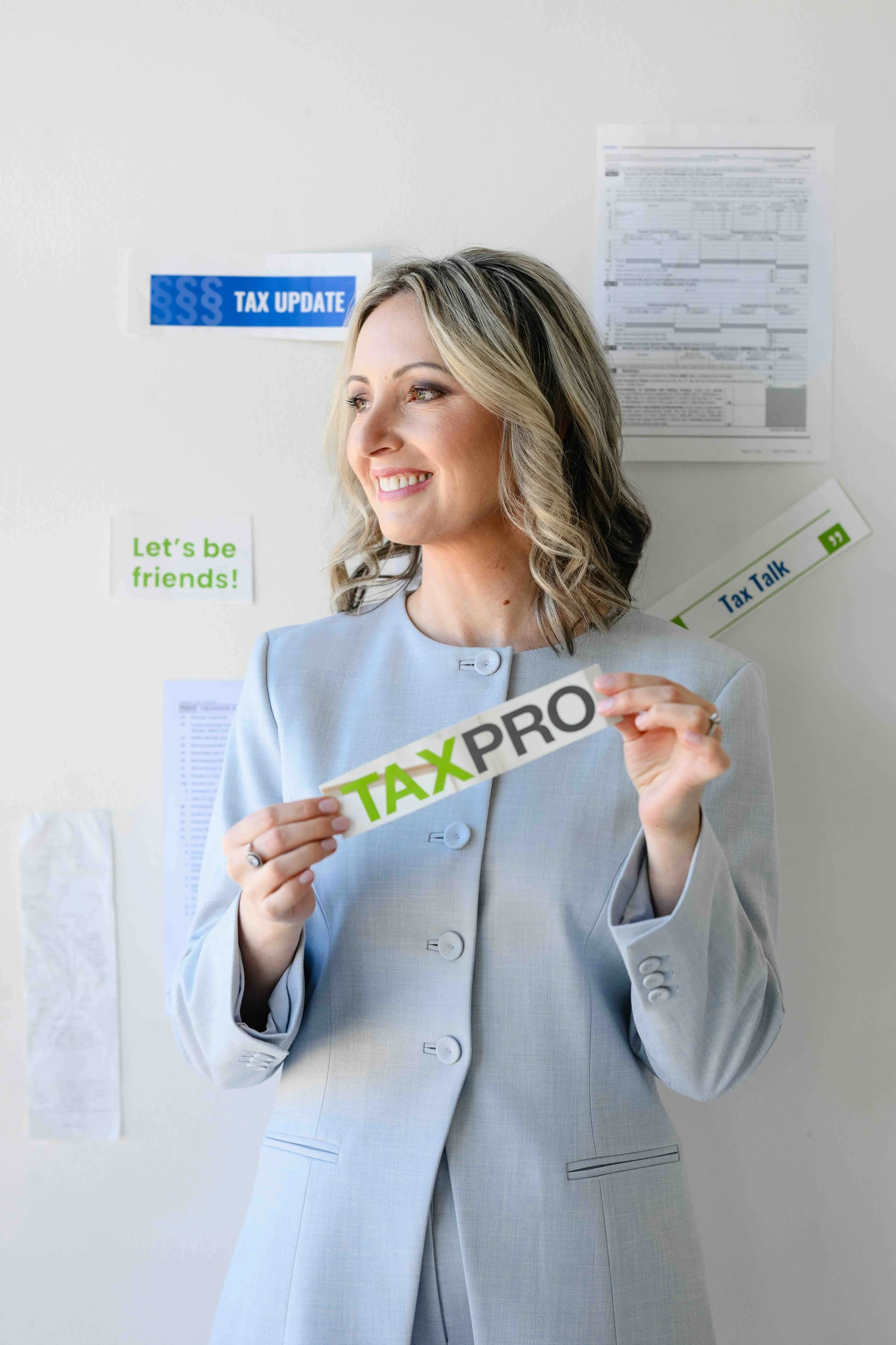 Woman in a light gray suit holding a sign that says 'TAX PRO' standing in front of a wall with documents including a blue 'TAX UPDATE' sign and a 'Let's be friends!' sticker.