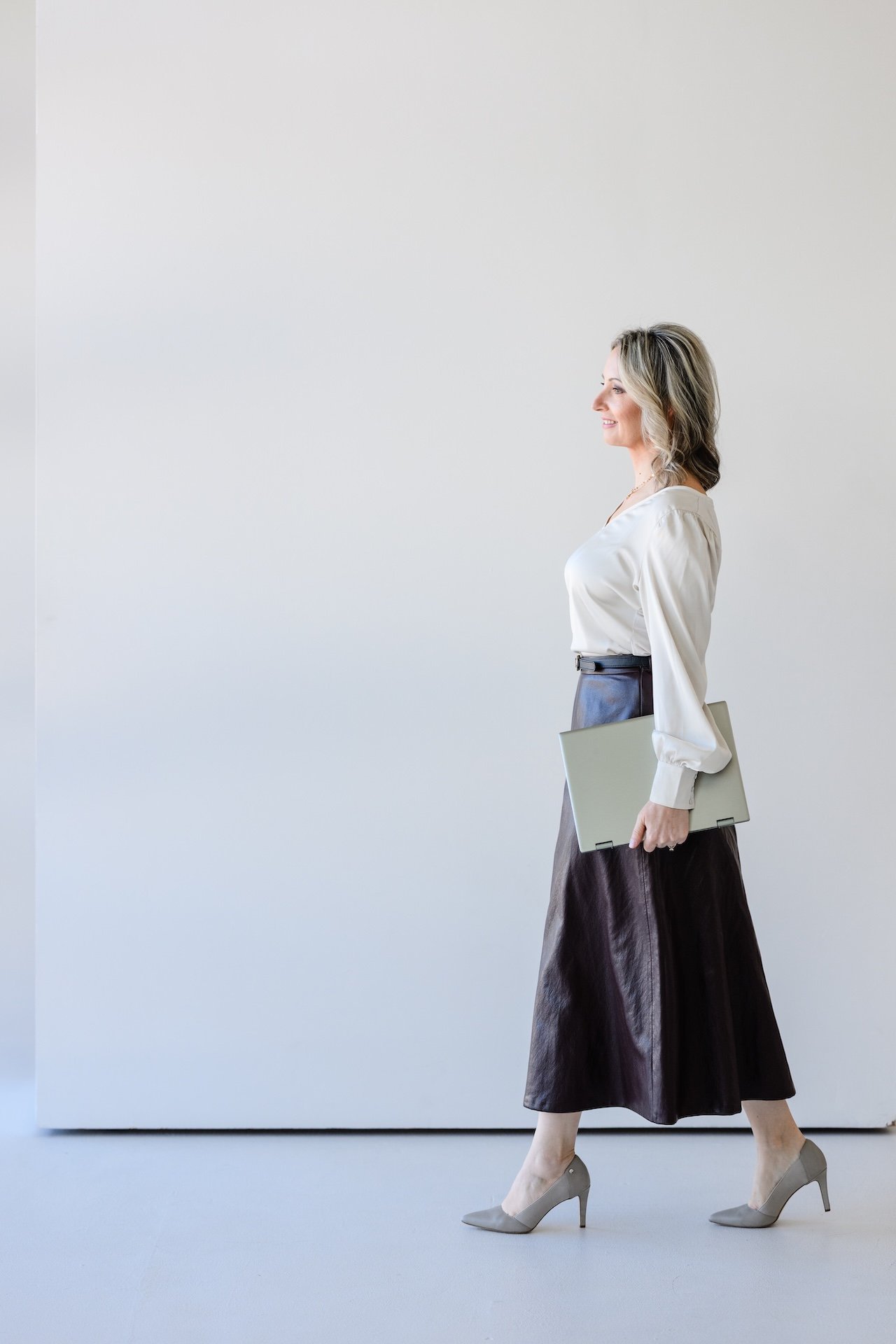 A woman walking in profile holding a laptop, wearing a white blouse, dark skirt, and high heels, with a neutral minimalist background.