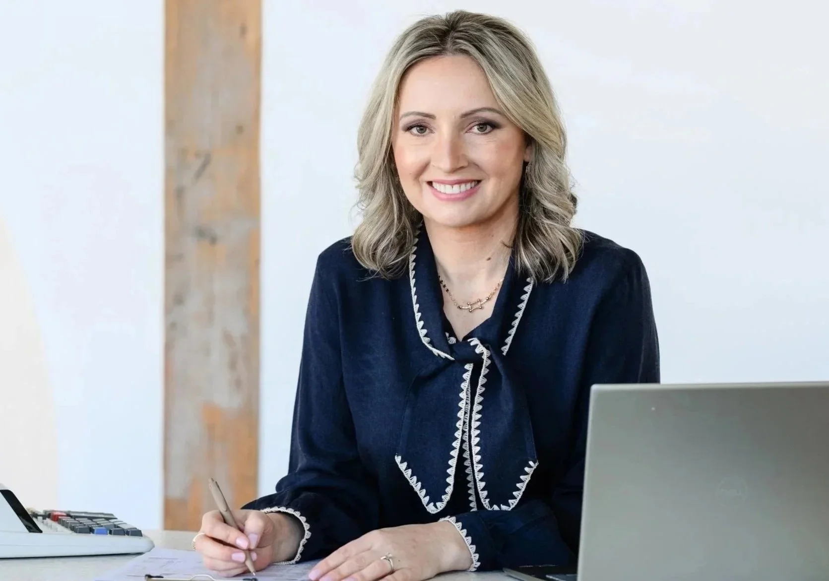 A woman with shoulder-length gray hair smiling at the camera, sitting at a desk with a laptop, writing on a notepad, with a wooden beam in the background.