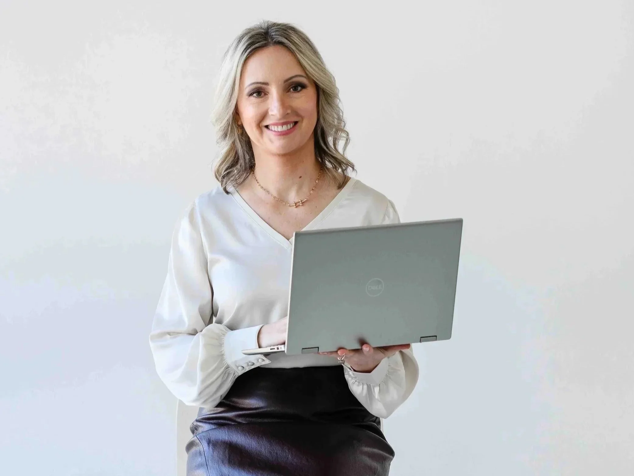 A woman with blonde hair holds a silver laptop, wearing a white blouse and a black skirt, standing against a plain white background.