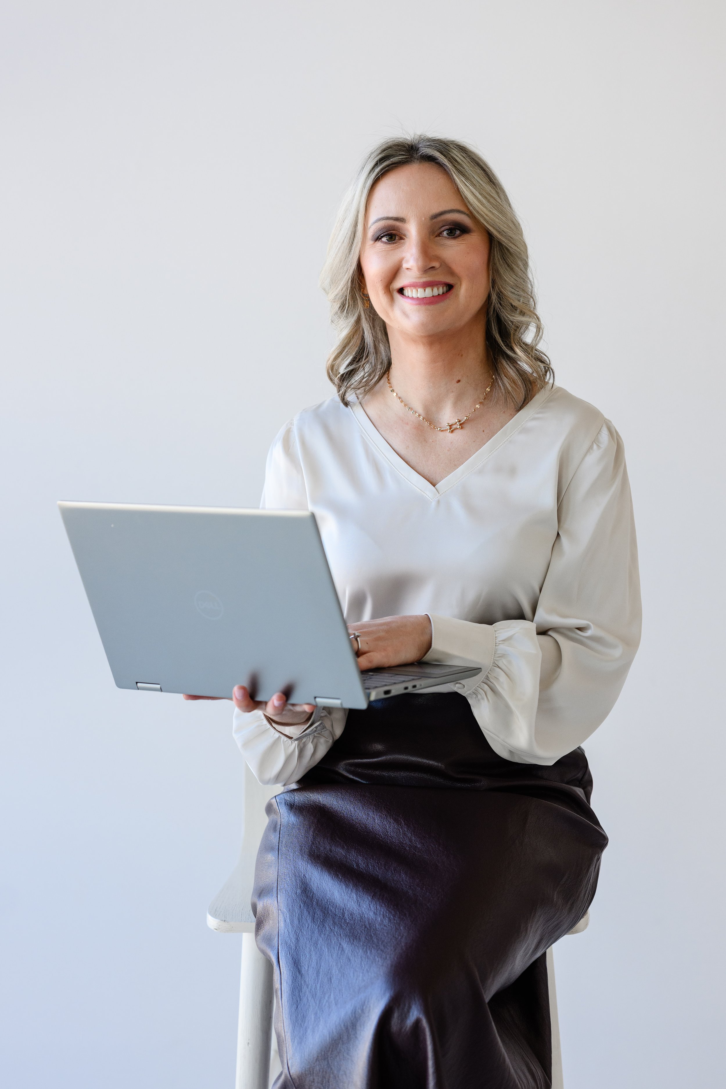 A smiling woman with blonde hair, wearing a cream-colored blouse and a dark satin skirt, sitting on a white chair while holding a silver laptop in her lap against a plain white background.
