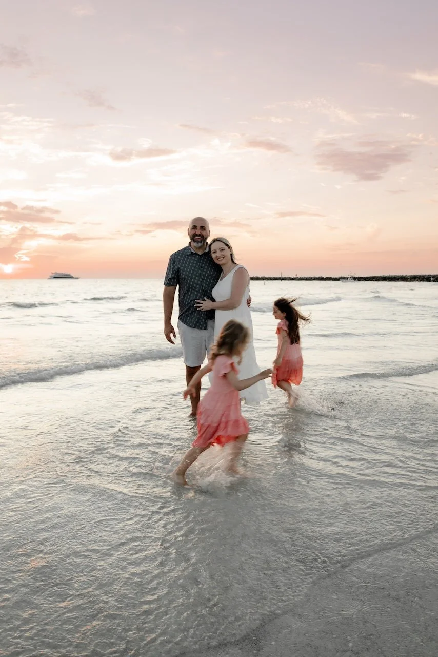 A family of four enjoying a day at the beach during sunset, with two young girls playing in the water and their parents standing together, smiling.
