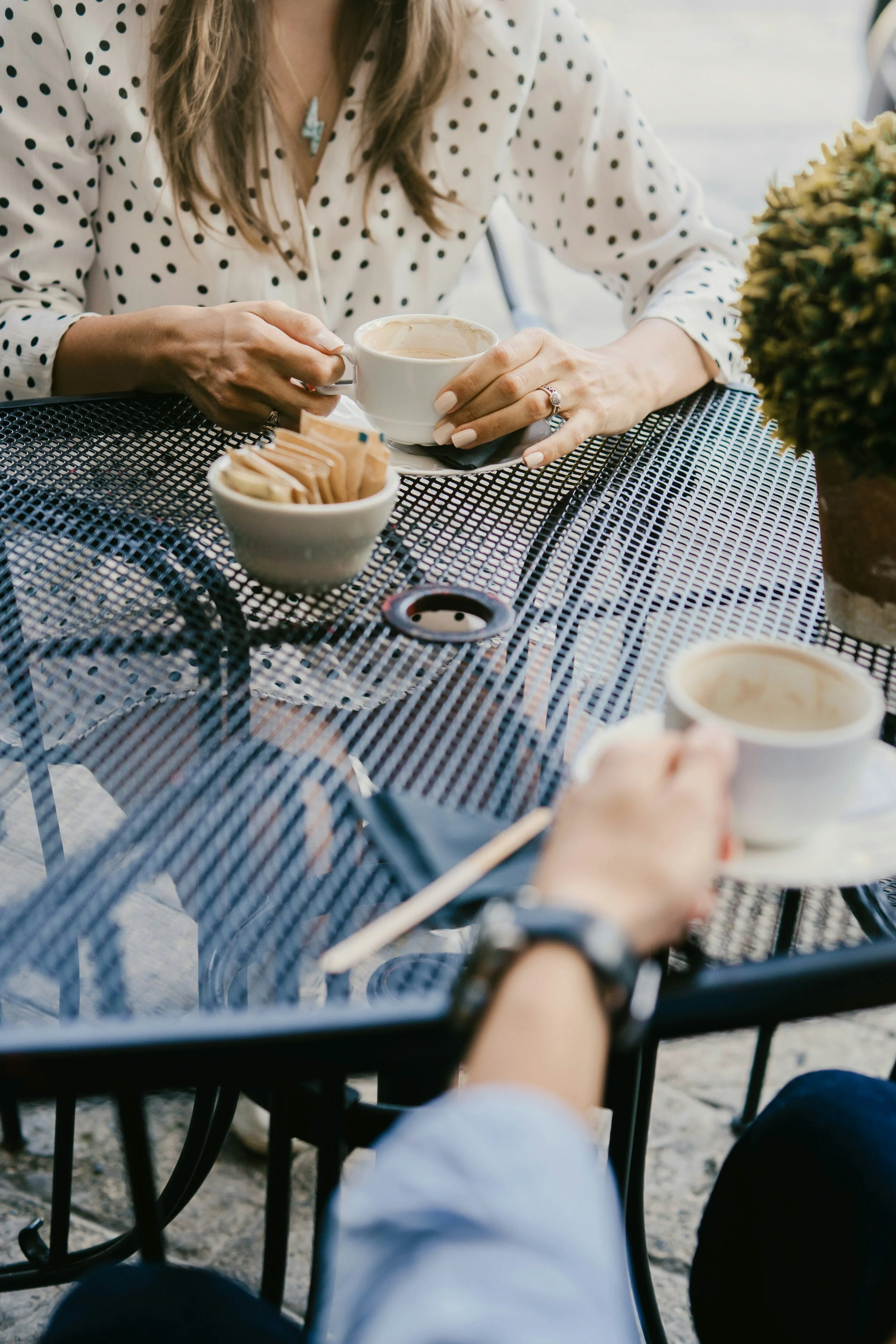 A woman with long hair wearing a polka-dot blouse and a lightning bolt necklace sits at a black metal outdoor table, holding a white coffee cup. There are small bowls with sugar packets and creamer on the table, with a blurry background.