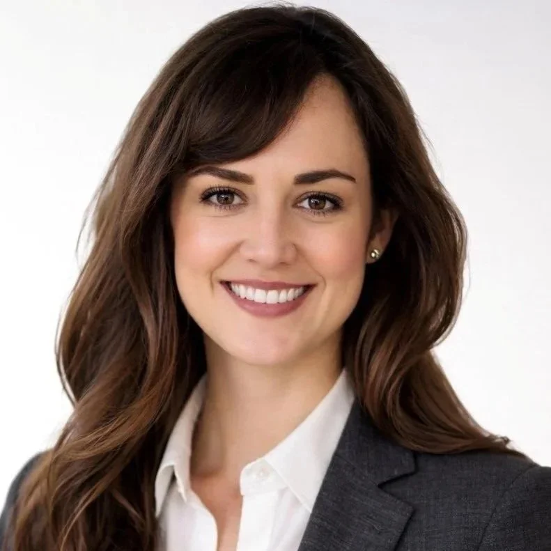 Professional woman with long brown hair, smiling, wearing a dark blazer and white shirt against a plain background.
