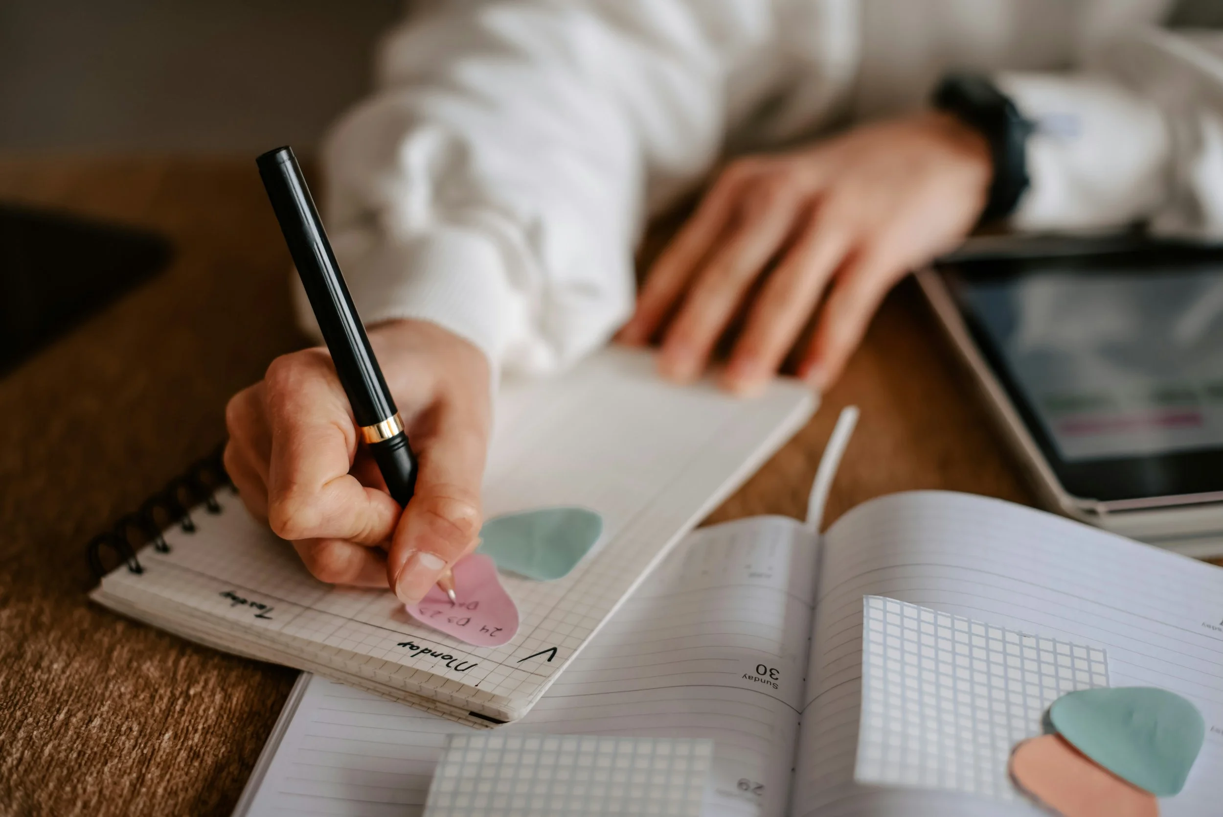 A person is writing in a spiral notebook with a black pen while sitting at a wooden desk. The person is wearing a white long-sleeve sweater and a black watch. Various colored paper circles and a pen are also on the open notebook, which has handwritten notes and a small graph.