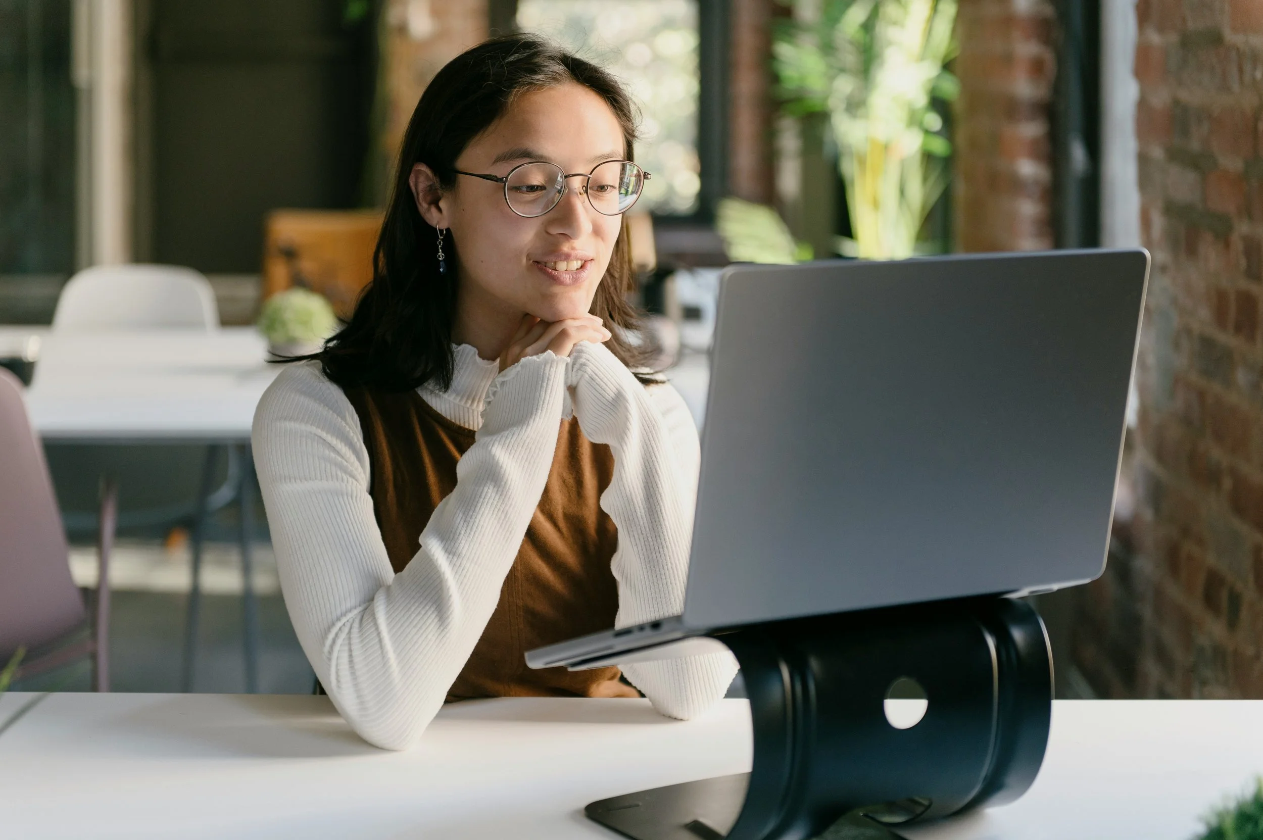 A young woman with glasses and earrings working on a laptop at a white table in a bright, modern workspace with large windows and greenery outside.