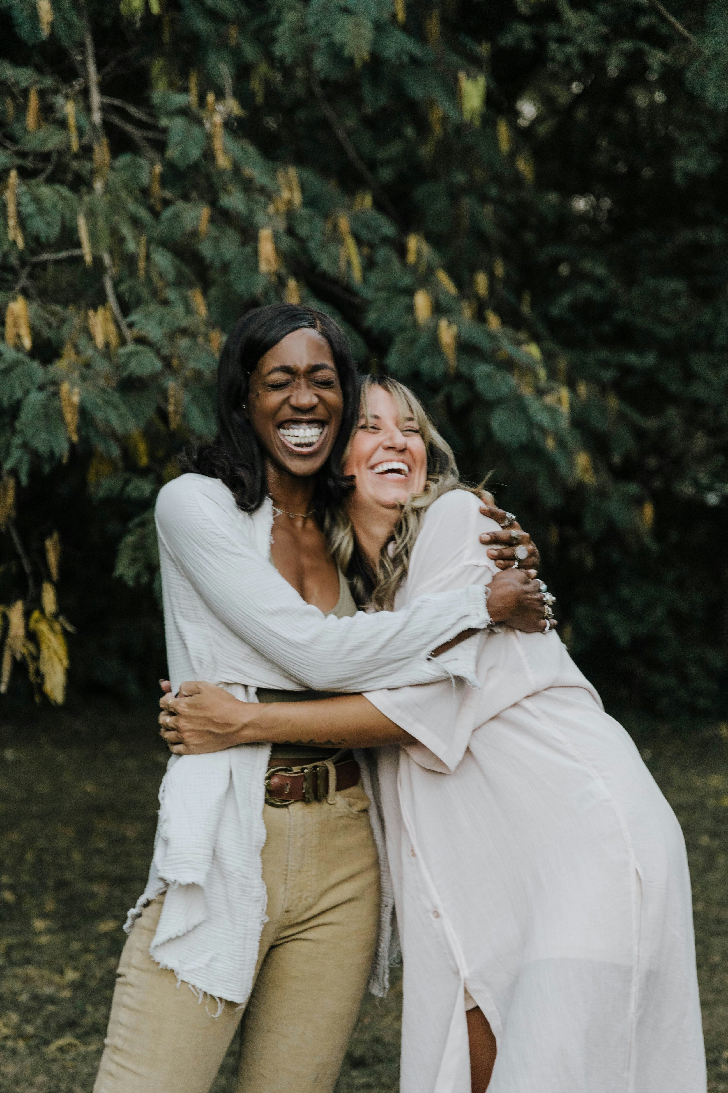 Two women are hugging and smiling outdoors with a green leafy background.
