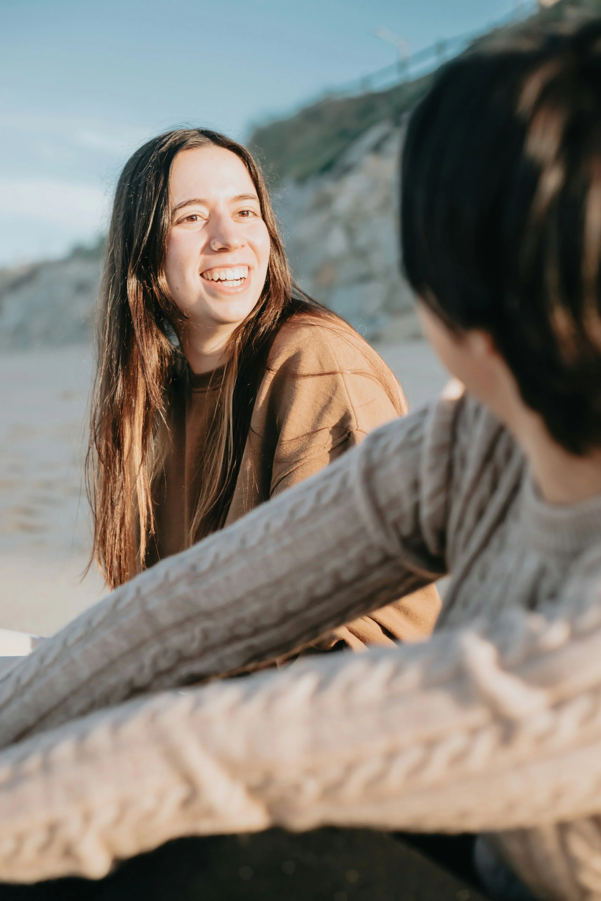 A woman with long brown hair, wearing a nose ring and a brown shirt, smiling and looking at a person with dark hair, on a beach with rocky cliffs and a blue sky in the background.