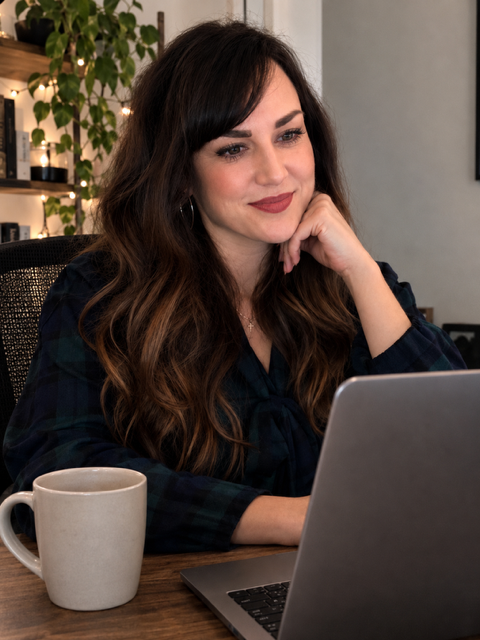 Woman with long brown hair and dark lipstick working on a laptop, sitting at a desk with a coffee mug, in a cozy indoor setting with plants and decorative lights.