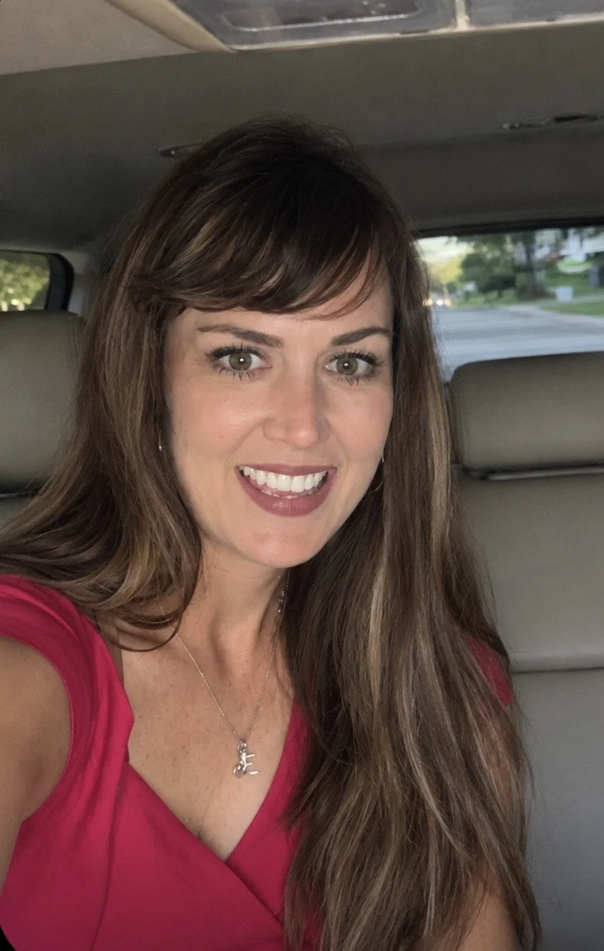 A woman with long brown hair smiling and wearing a red top, sitting inside a vehicle.