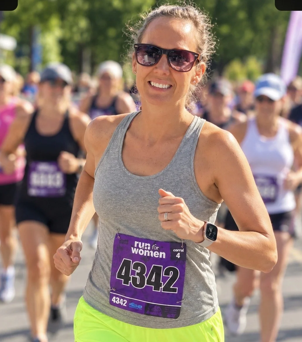 A smiling woman wearing sunglasses and a gray tank top running in a race with a purple bib labeled 4342, surrounded by other runners.