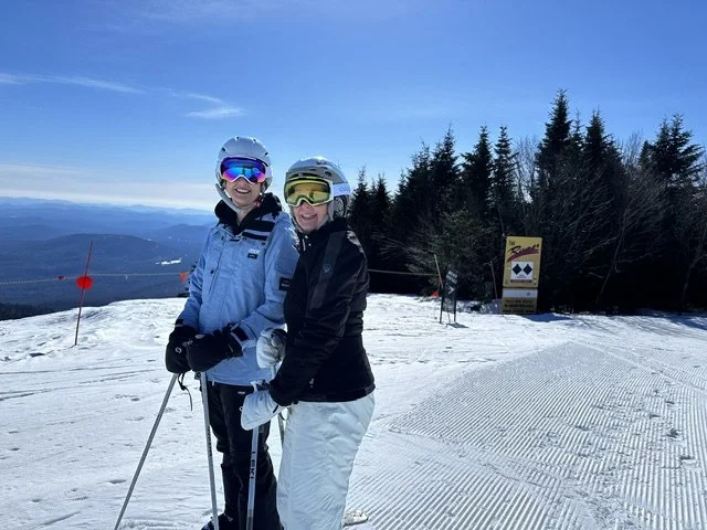 Two people wearing ski helmets and goggles standing on a snowy ski slope with trees and a sign in the background.