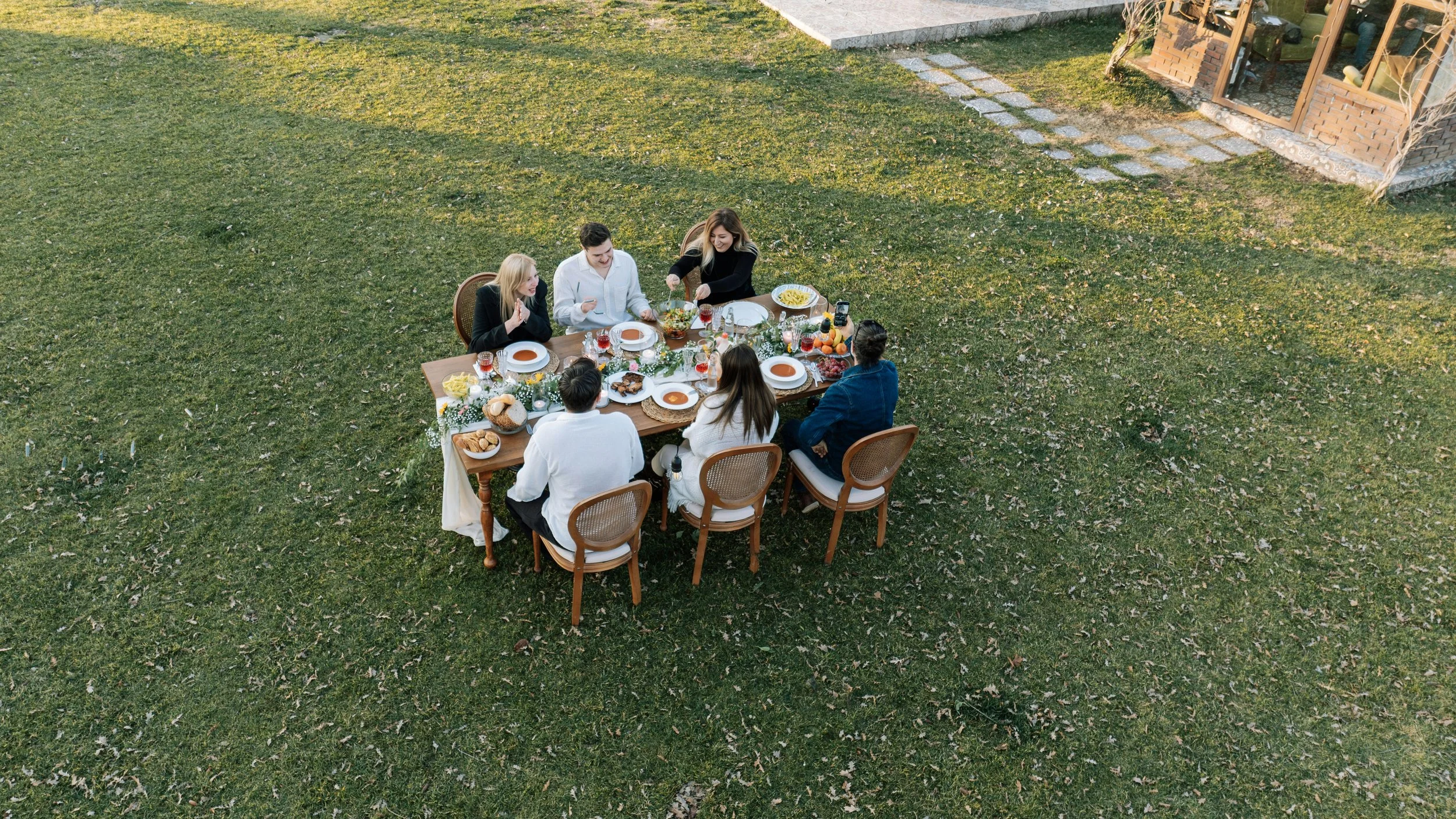 A group of six people sharing a meal at a long outdoor table on a grassy yard, with a house in the background.