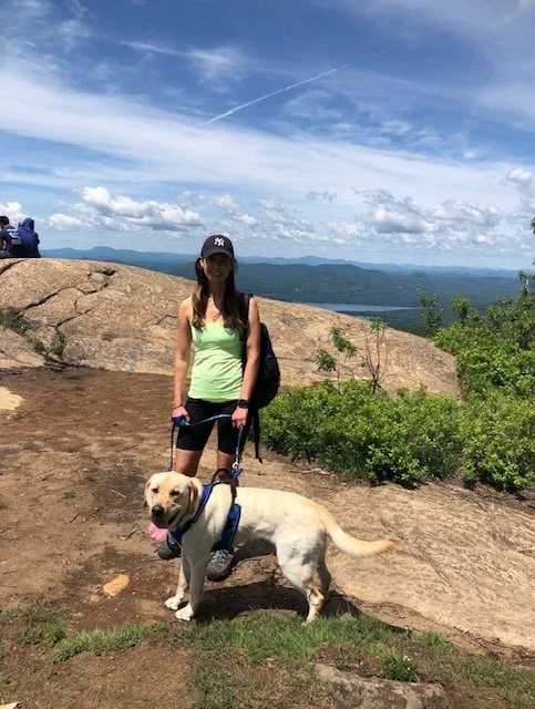 A woman with a backpack and a cap standing on a rocky hilltop with a Labrador Retriever dog in front of her. There are green bushes and a view of mountains and a lake in the background under a partly cloudy sky.