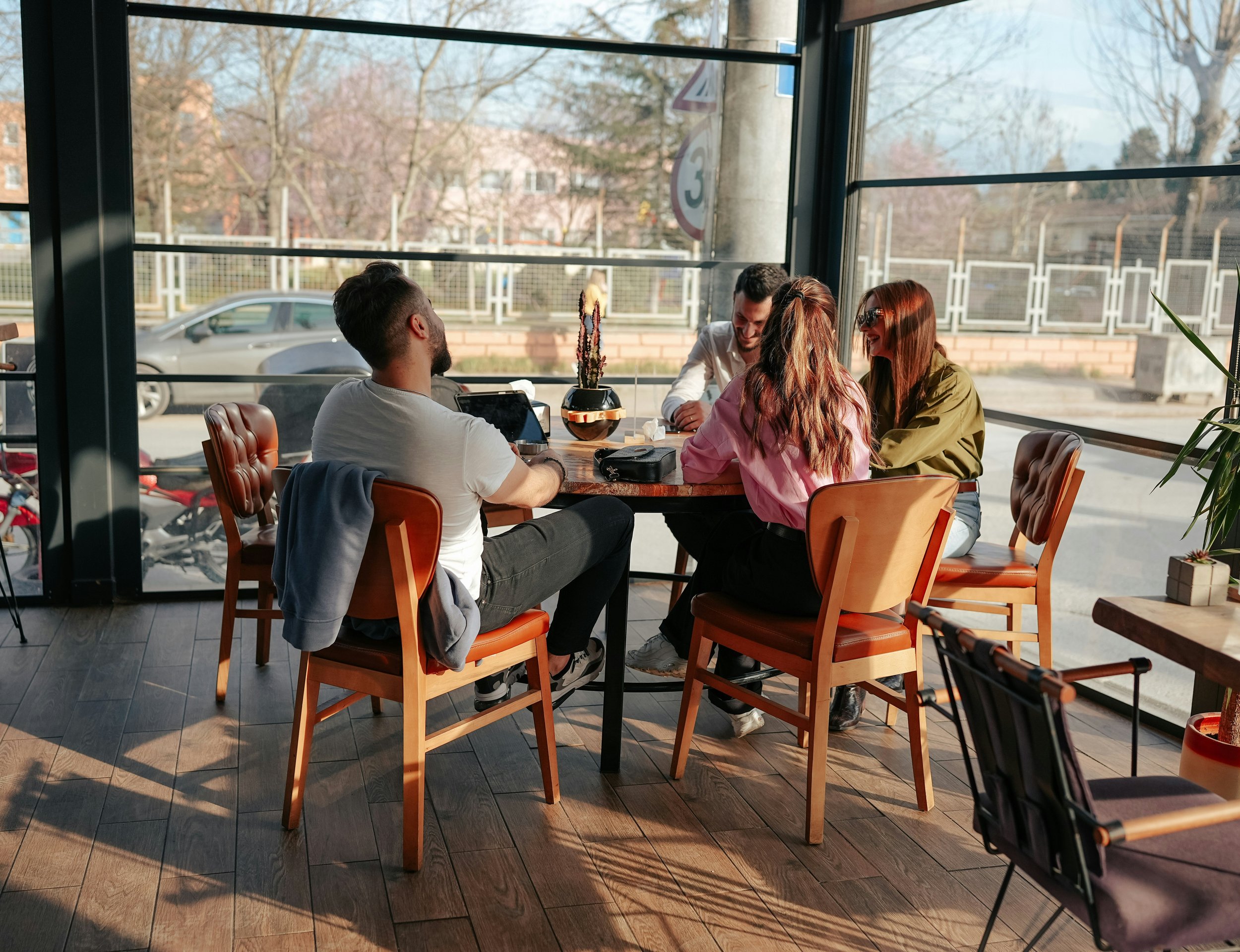 Four friends sitting around a wooden table in a restaurant with large windows, enjoying sunlight and laughter, with trees and parked cars outside.