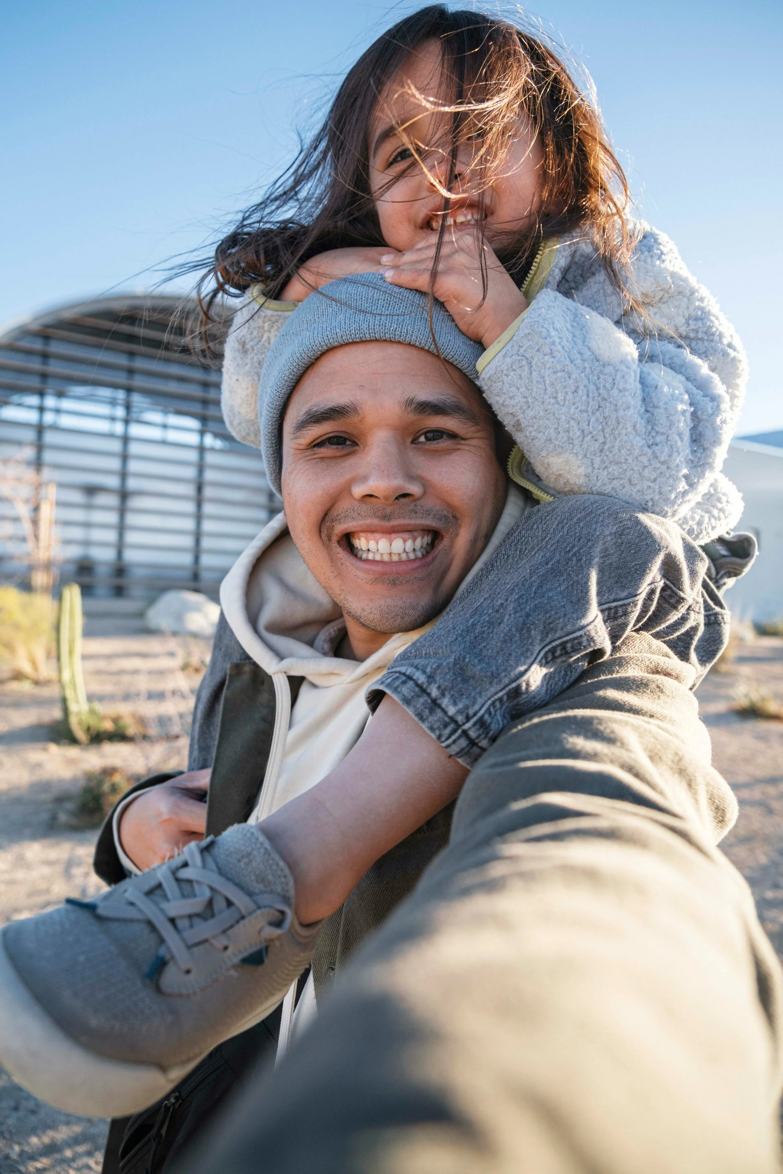 Smiling man with a child on his shoulders, outdoors on a sunny day, with modern building and cacti in the background.