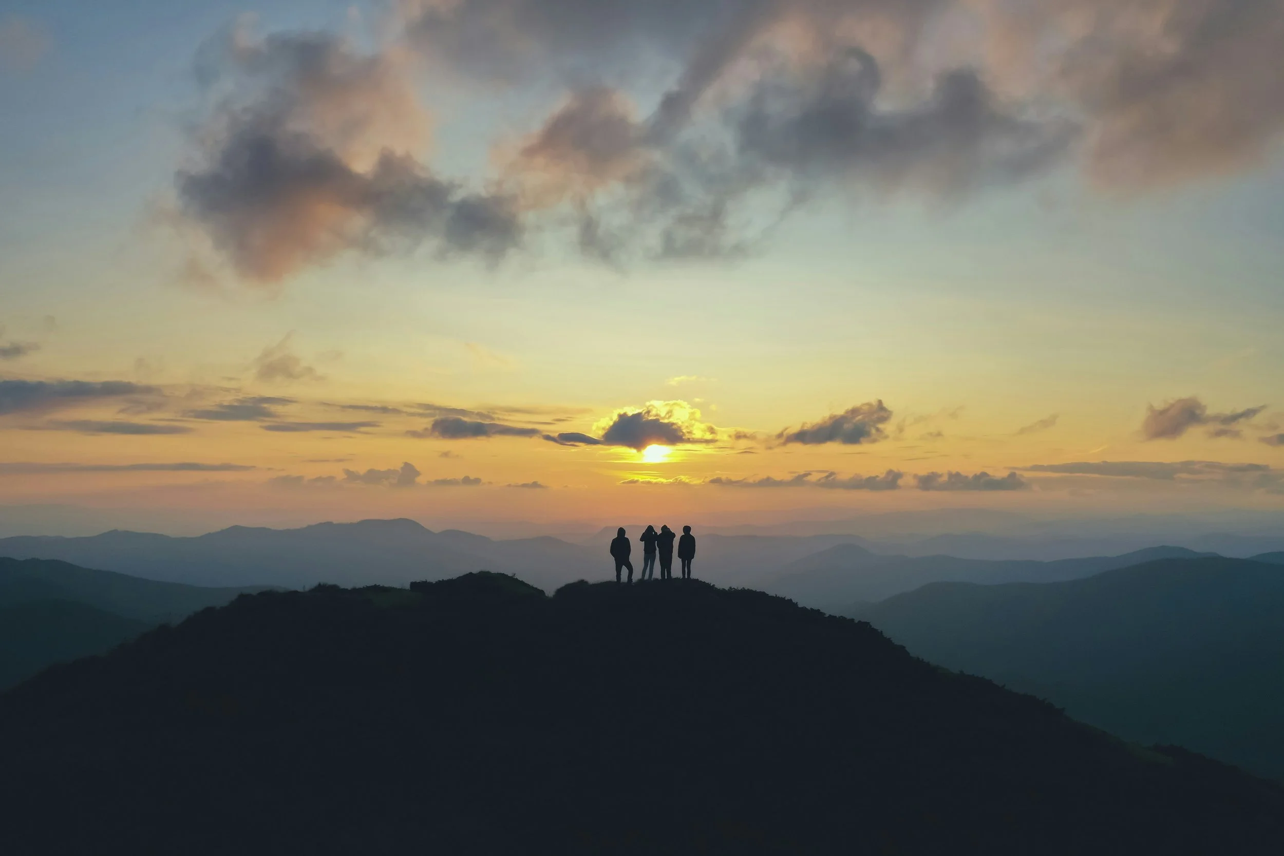 Three people standing on a hilltop, silhouetted against a sunset sky with clouds and mountain ranges in the distance.