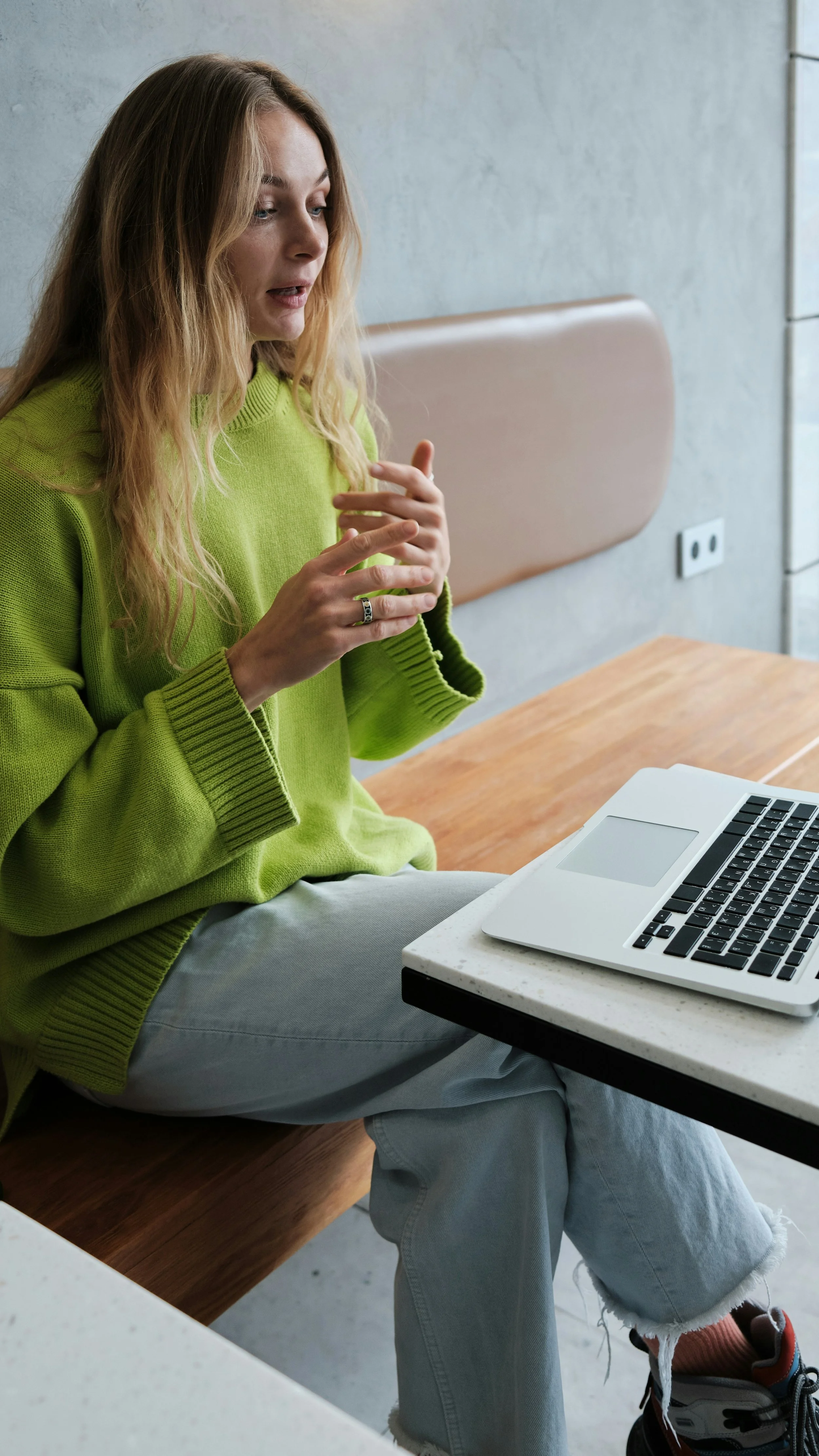 A young woman sitting at a table with a laptop, wearing a lime green sweater and light blue jeans, in a modern space.