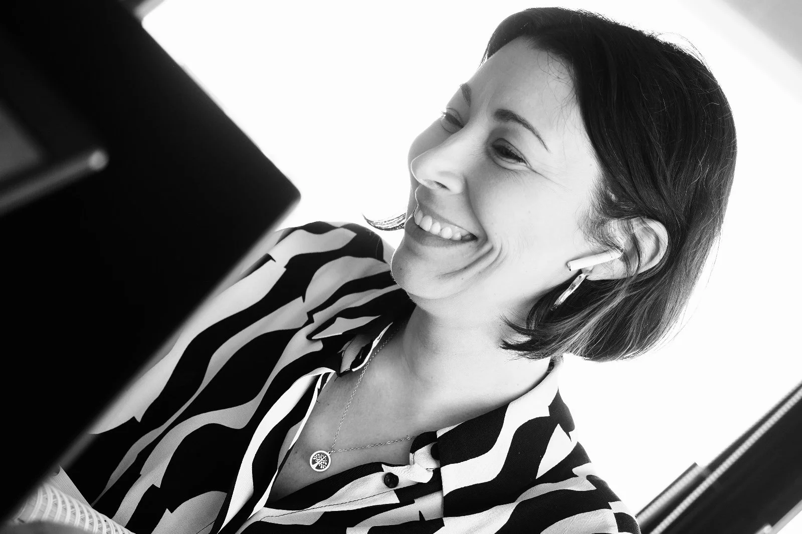 Mulher sorridente ajustando seus fones de ouvido, de cabelo curto e liso, usando camisa de estampa listrada, em um ambiente bem iluminado.