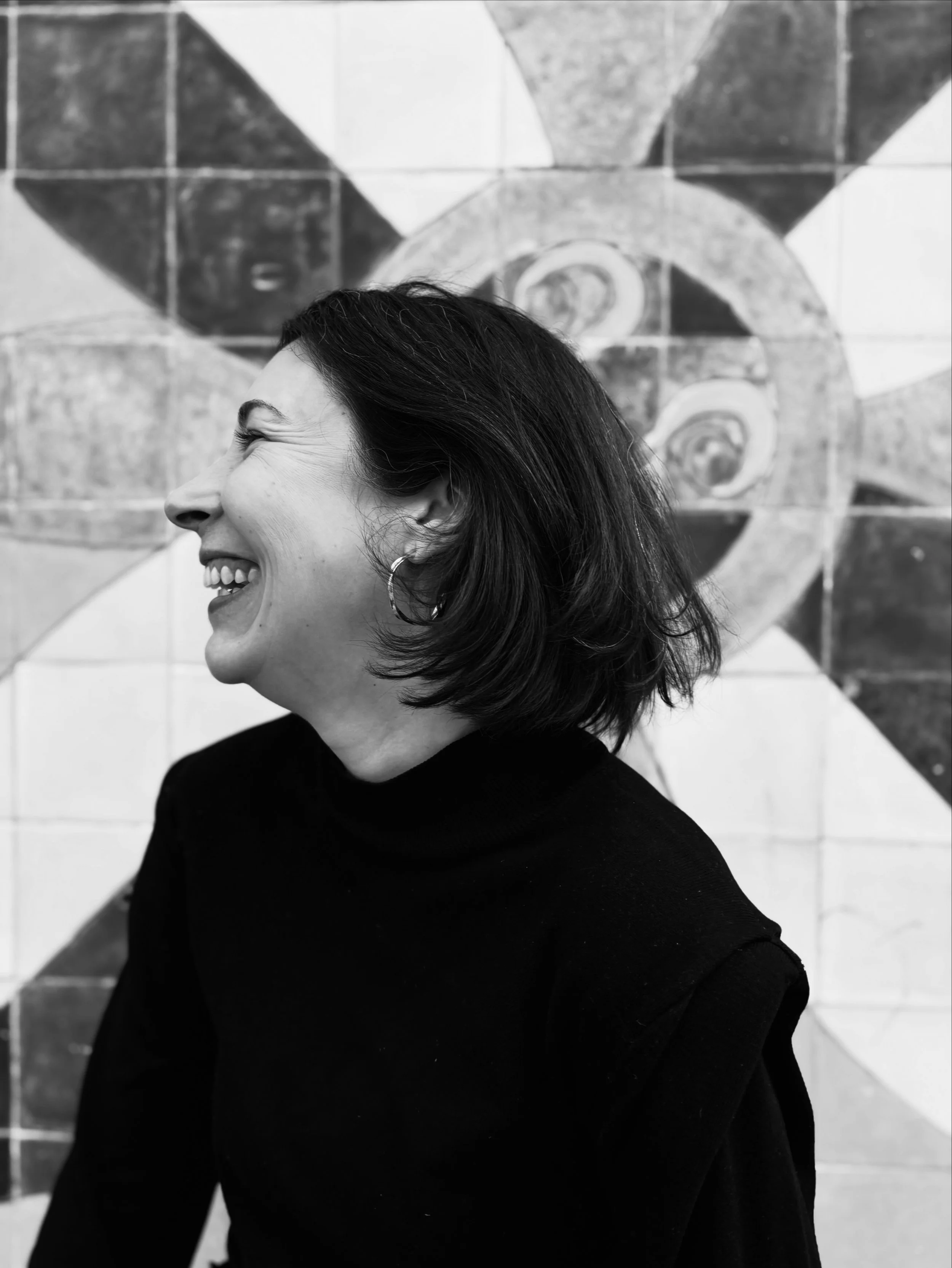 Retrato de uma mulher sorridente com cabelo escuro e cacheado, usando brincos de argola preta, em frente a um mural de azulejos com padrão geométrico em tons de preto e branco.