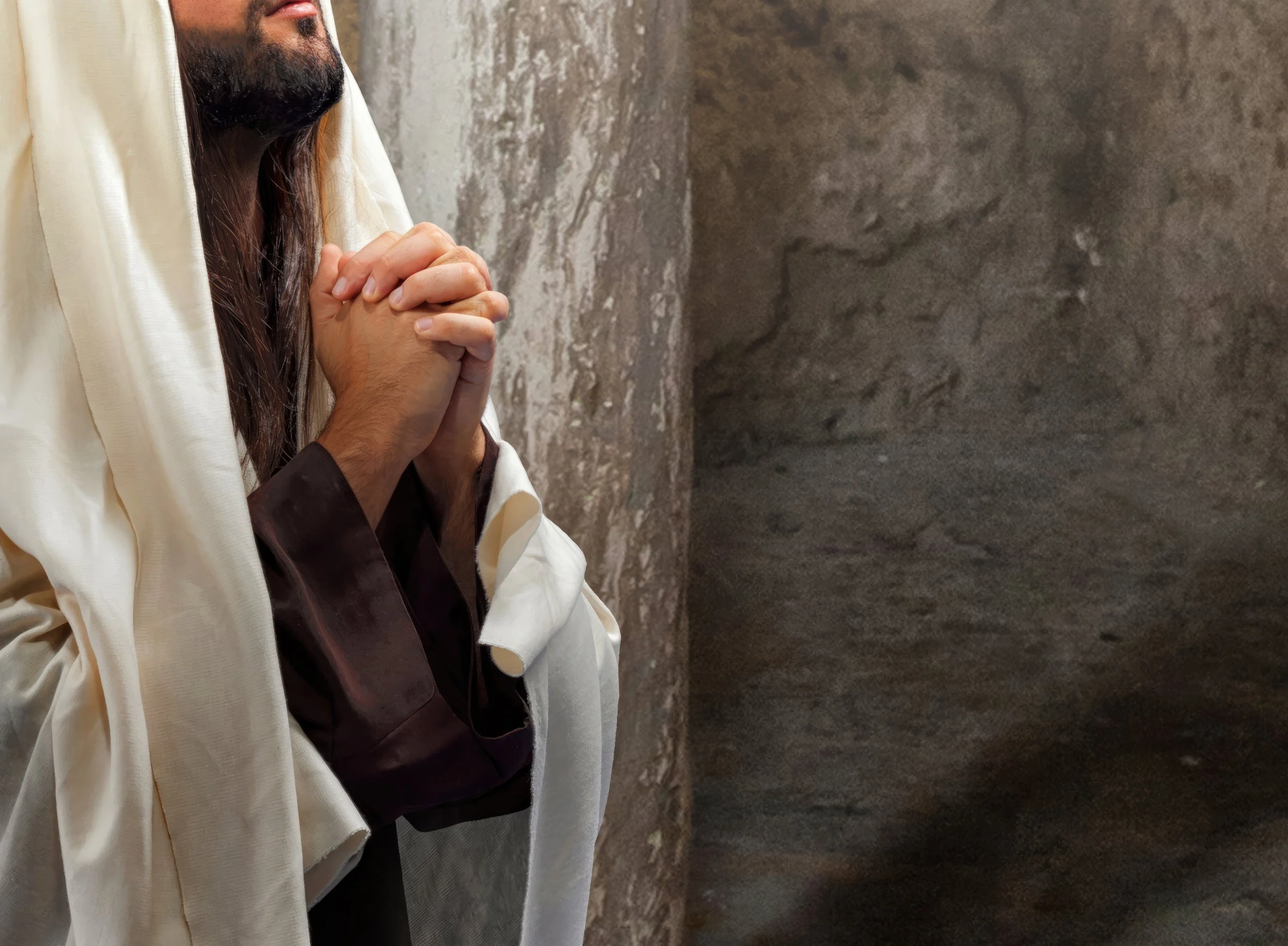 A person dressed in robes, possibly depicting a religious figure, is seen with hands clasped in prayer in front of a textured stone wall.