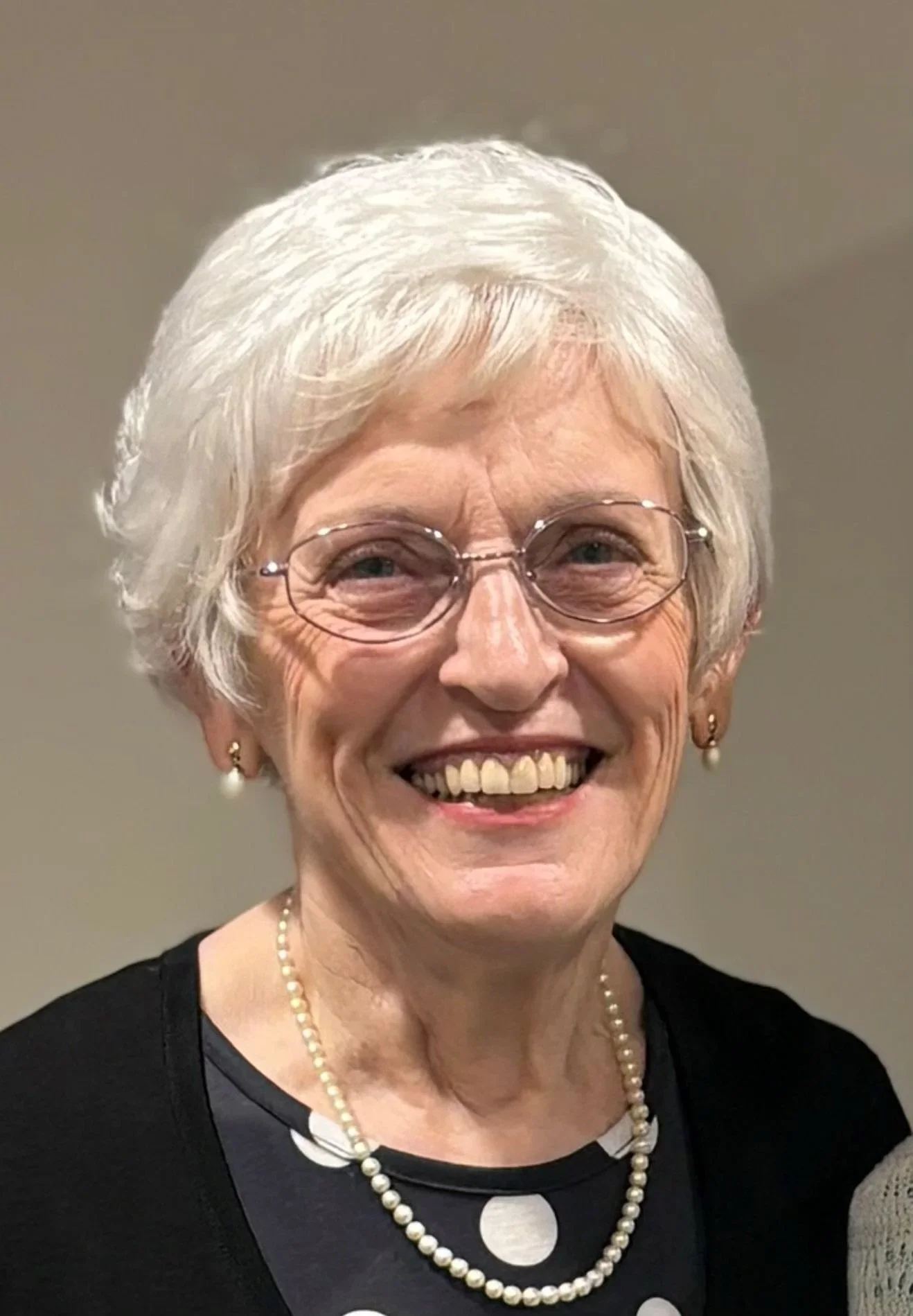 A smiling elderly woman with short white hair, wearing glasses, pearl earrings, a pearl necklace, and a polka dot dress with a black cardigan, standing against a plain background.