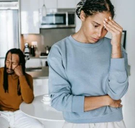Two women appear upset in a kitchen, one is holding her head with her hand, and the other is sitting at a table with her hand on her forehead.