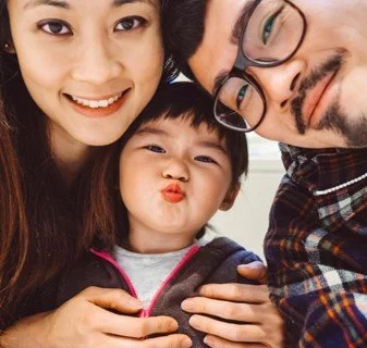 A happy family of three taking a selfie together indoors, with a woman on the left, a man on the right, and a young girl in the middle making a duck face.