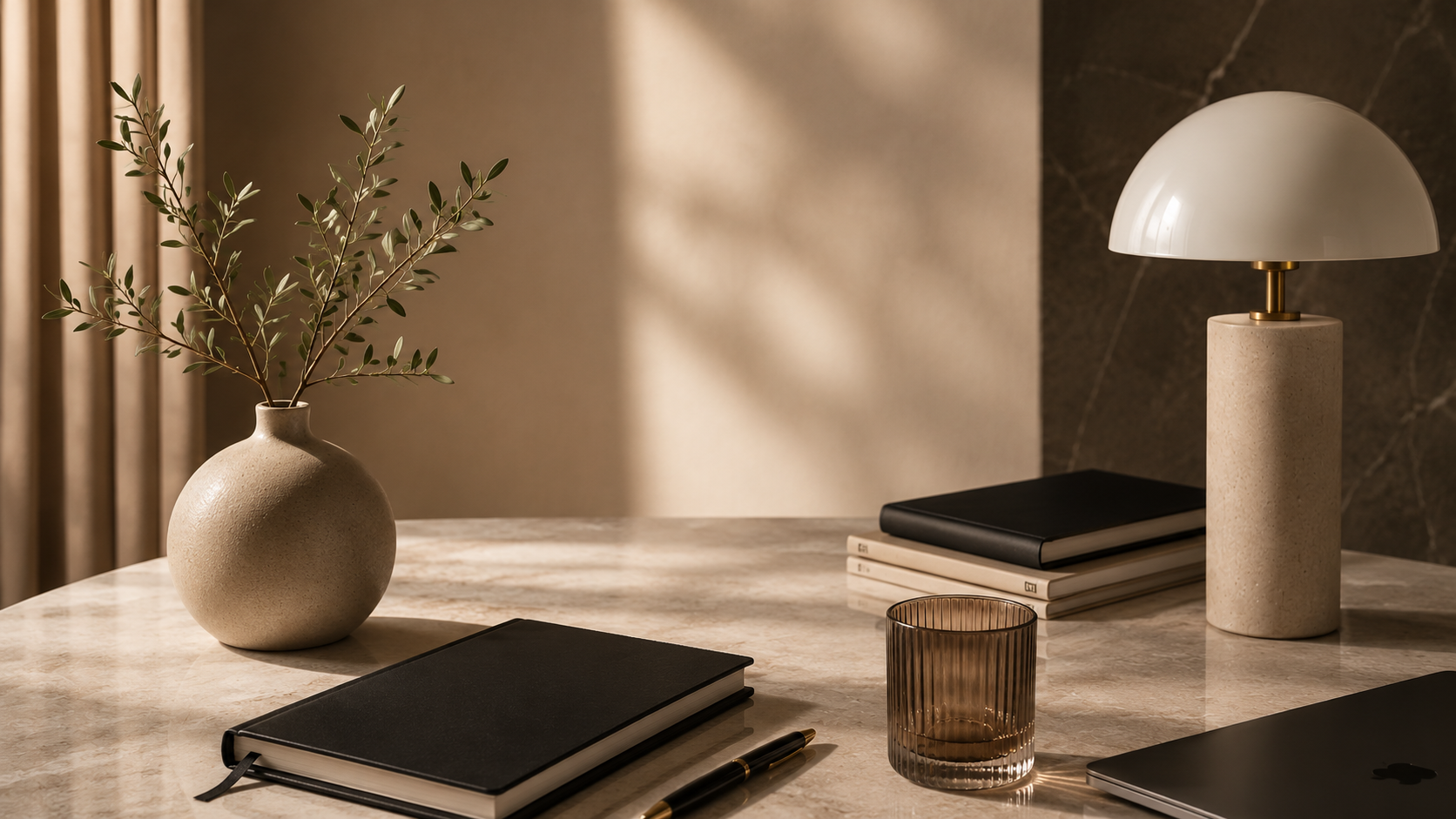 Une table avec un vase en céramique contenant des branches vertes, une lampe de bureau en céramique avec abat-jour blanc, un carnet noir, une pile de livres, un verre teinté, un stylo or et un ordinateur portable.