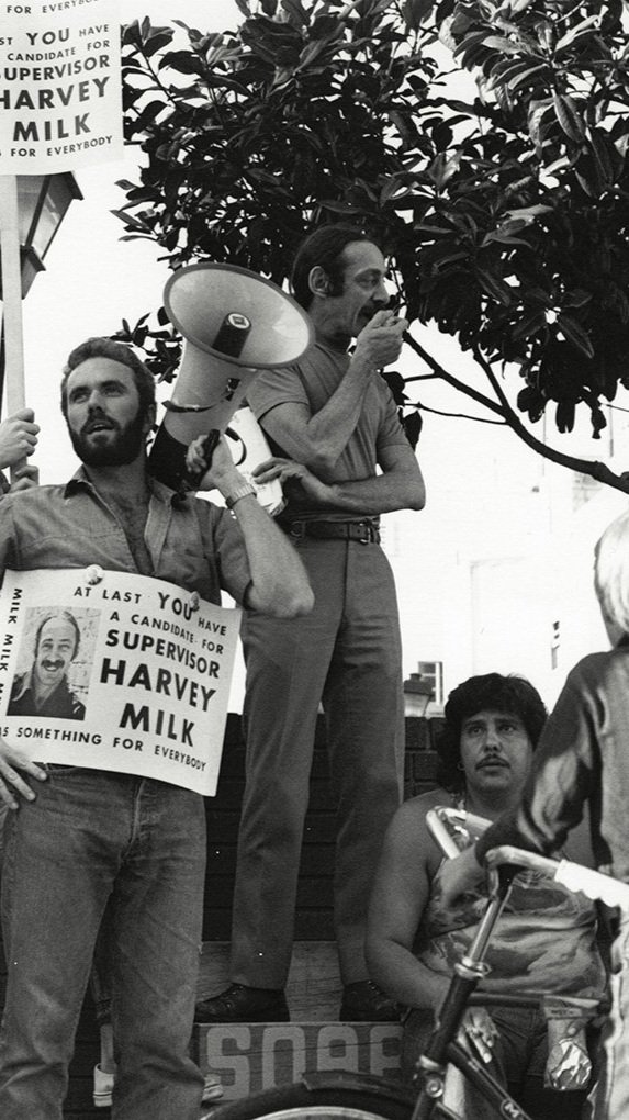 Harvey Milk campaigning for Supervisor. A picket sign reads "At last YOU have a candidate for supervisor. Harvey Milk, something for everybody."