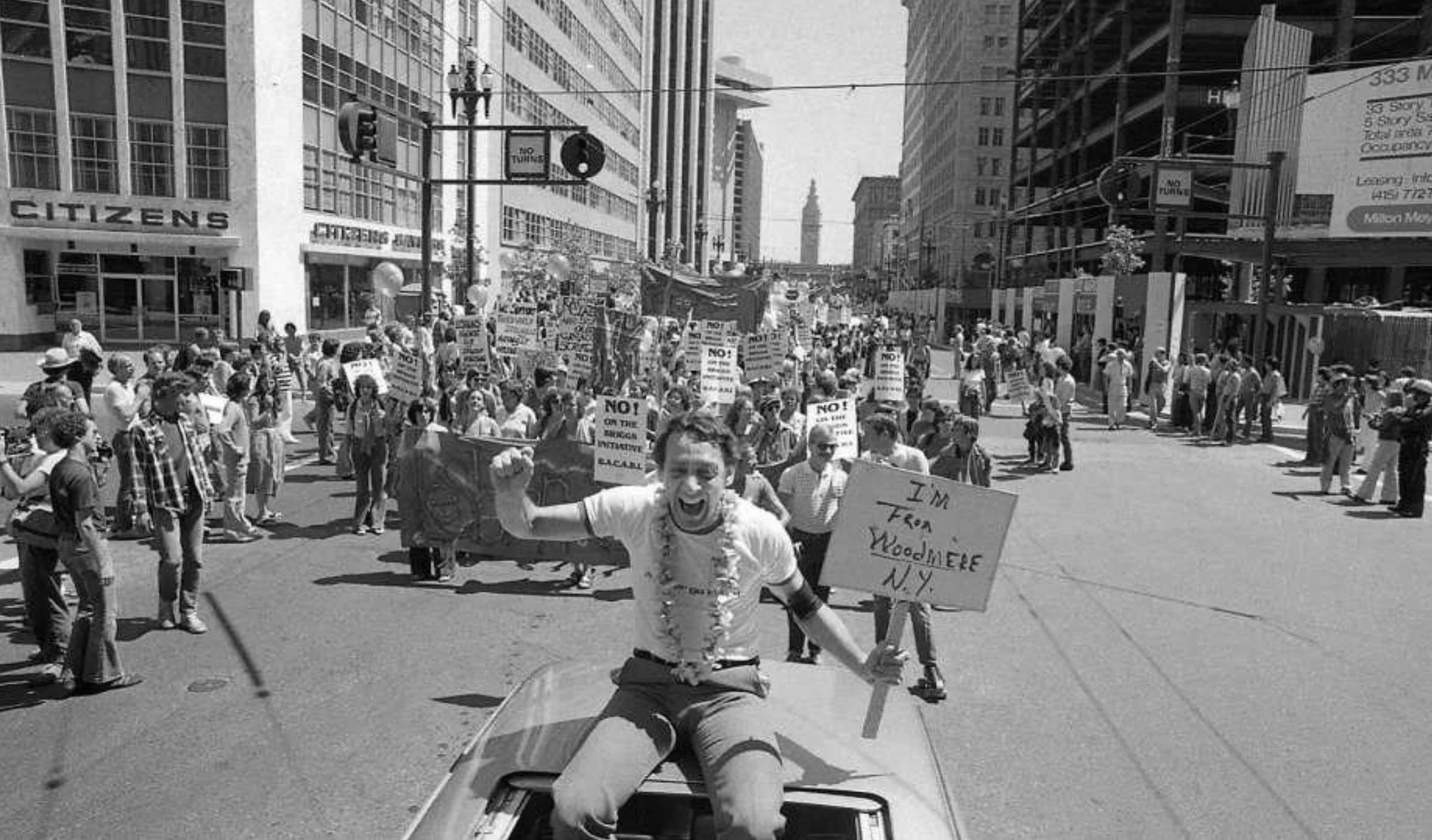 Black and white photo of Harvey Milk wearing a flower garland in the back of a car. He's holding a sign reading, "I'm from Woodmere, N.Y."
