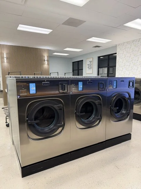 Three silver front-loading washing machines in a laundromat, with digital displays and control panels.