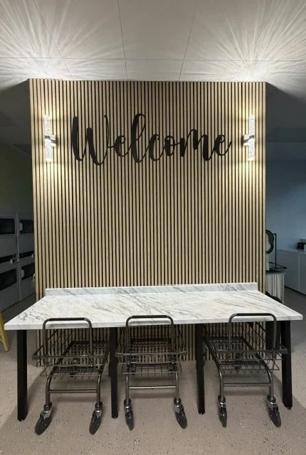 Interior of a room with a wooden slat wall, with the word 'Welcome' in cursive. A white marble table with four shopping carts lined up in front.
