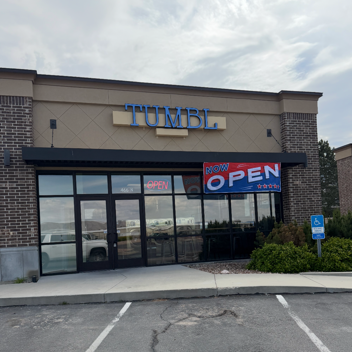 Facade of a store named TUMBL with a large 'Now Open' banner in front, and pink neon 'Open' sign inside, in a strip mall parking lot with reserved handicap parking sign.