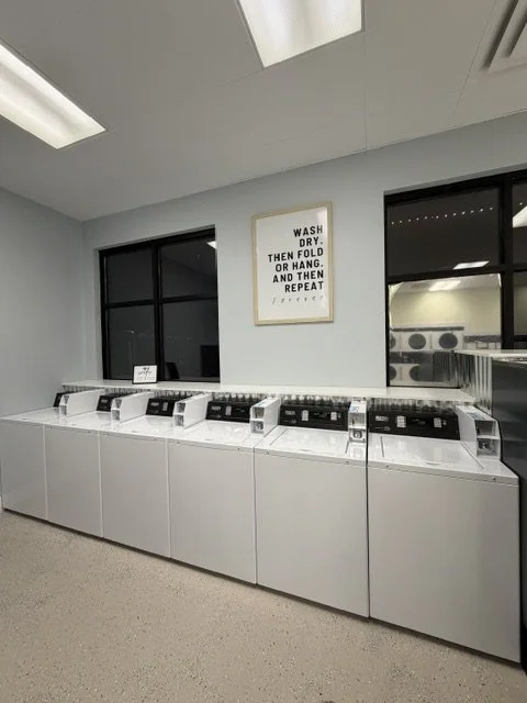 Row of six white and black laundry washing machines in a laundromat with two large windows and a framed sign on the wall