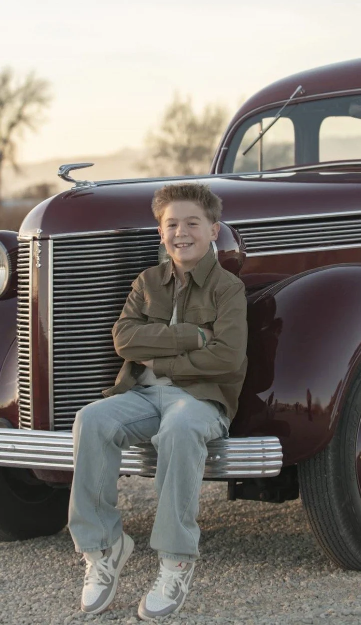 A boy sitting on the bumper of a vintage maroon car, smiling, outdoors during sunset, wearing a brown jacket, light jeans, and sneakers.