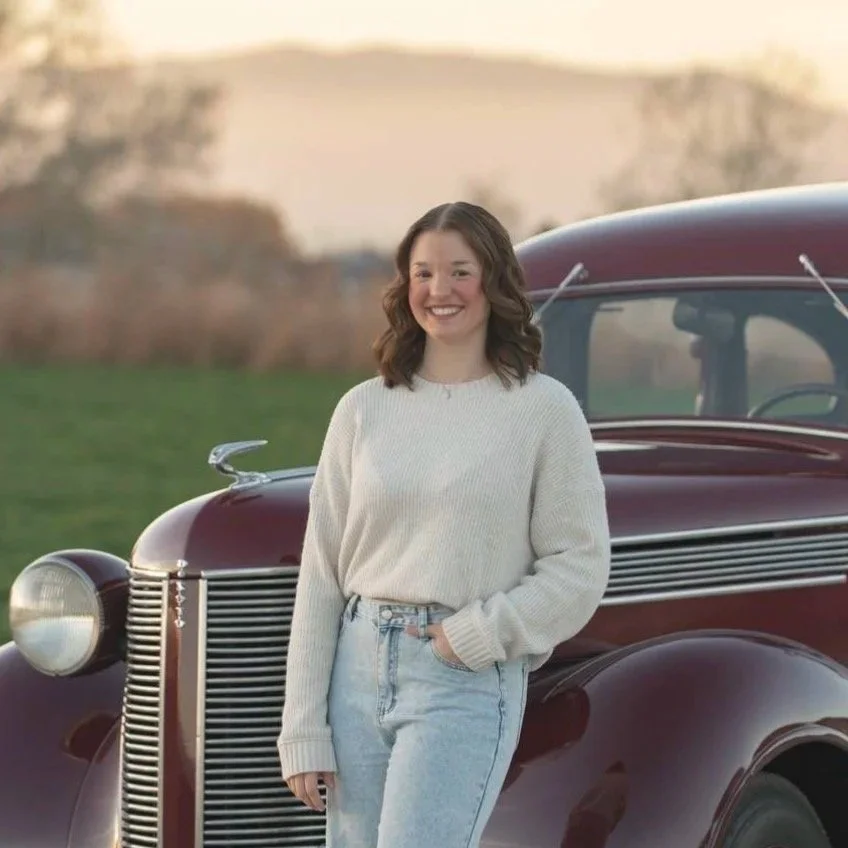 A young woman with shoulder-length wavy brown hair, wearing a cream sweater and light blue jeans, stands smiling next to a vintage maroon car outdoors during sunset.