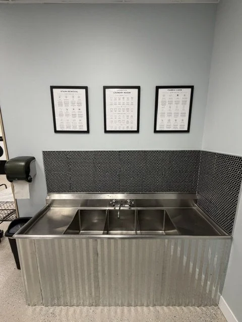 A stainless steel handwashing sink with a divided basin, mounted against a light gray wall with three framed signs above it. A black wall-mounted paper towel dispenser is on the left side of the sink.