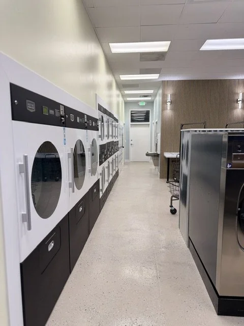 Laundry room with multiple washers and dryers along the left wall and stainless steel machines or counters on the right side, with a door at the end of the room.