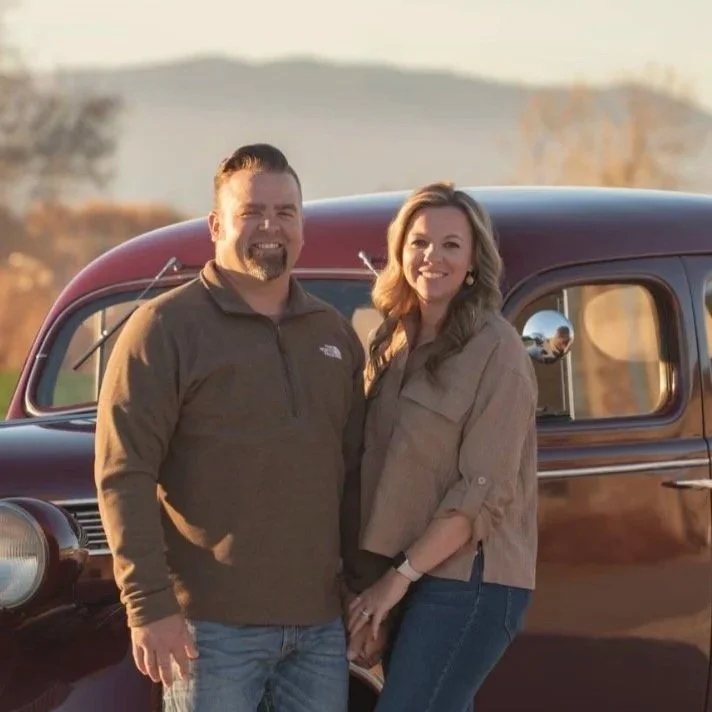 A smiling man and woman standing close together in front of a vintage car, holding hands outdoors during sunset, with a scenic landscape in the background.