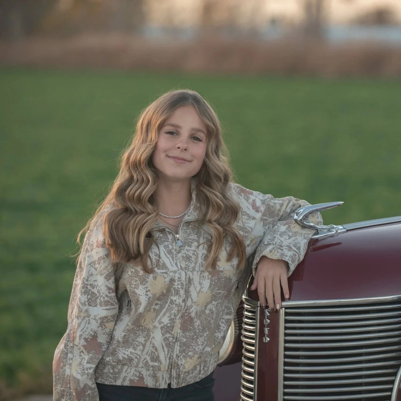 A young woman with wavy blonde hair smiling, standing outdoors in front of a green grassy area and a blurred background, leaning on a vintage red car.