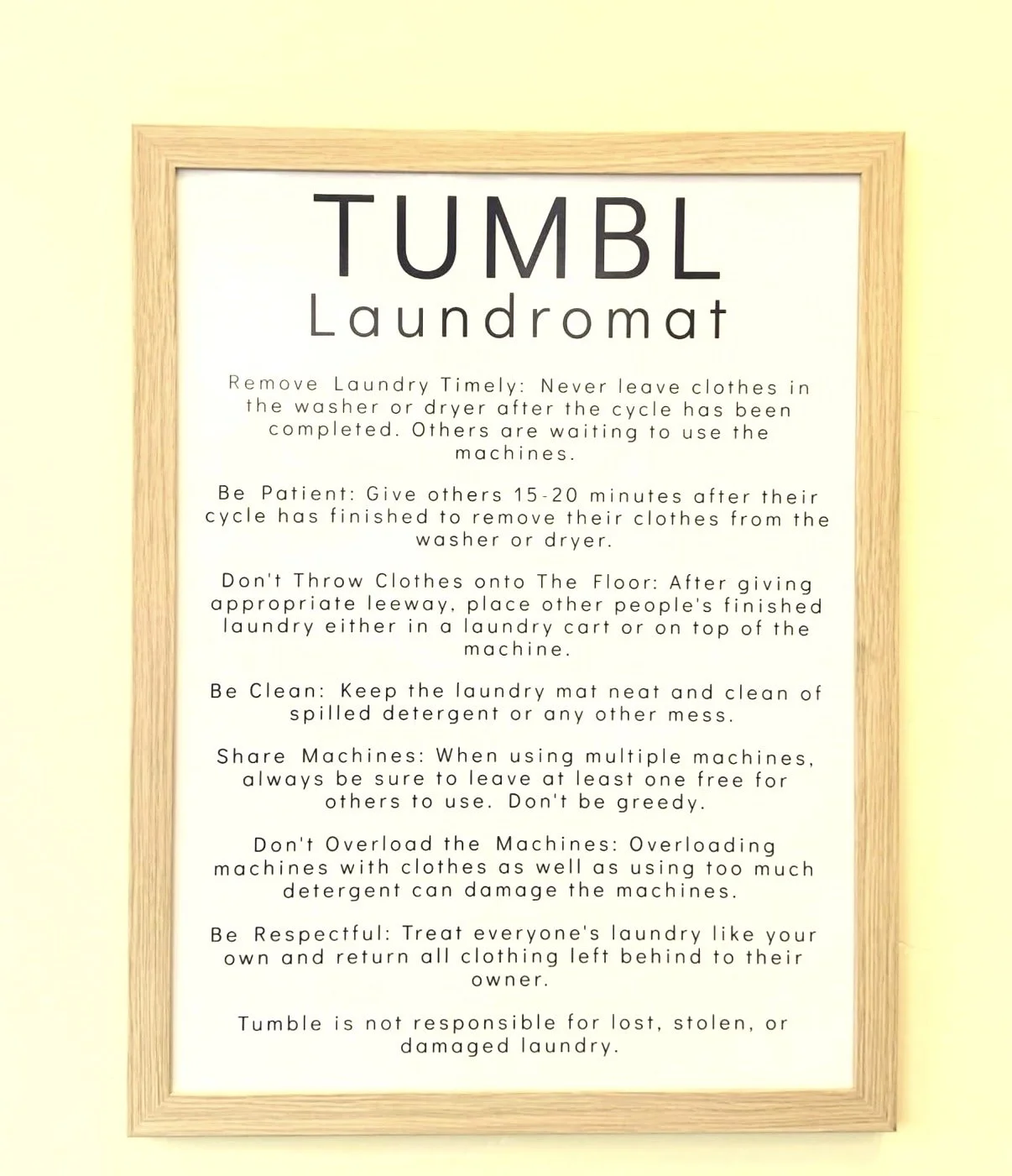 Instruction sign with laundry etiquette tips at the Tumble Laundromat, framed in wood, on a light-colored wall.