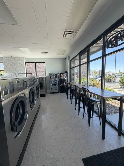 Empty laundromat with washing machines, vending machines, a row of chairs, and large windows showing a parking lot outside.