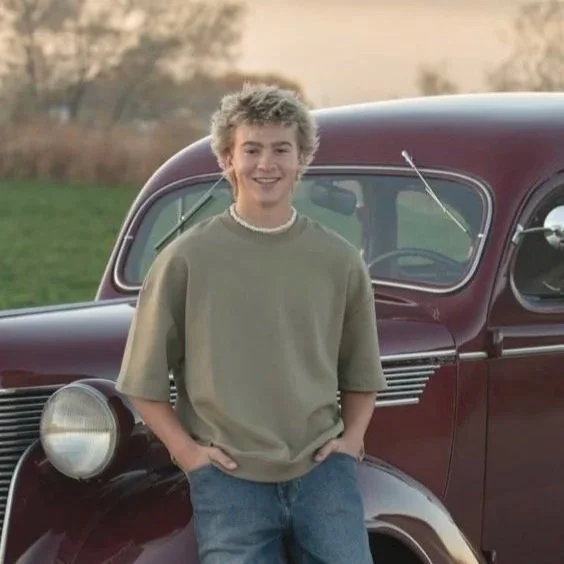 A young person with curly blonde hair smiling and leaning against the front of a vintage maroon car, outdoors on a grassy field with trees in the background.