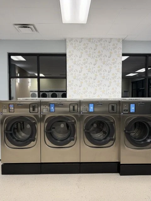 Four front-loading commercial washing machines in a laundromat. Two windows in the background, plain ceiling, and a column with a floral pattern.