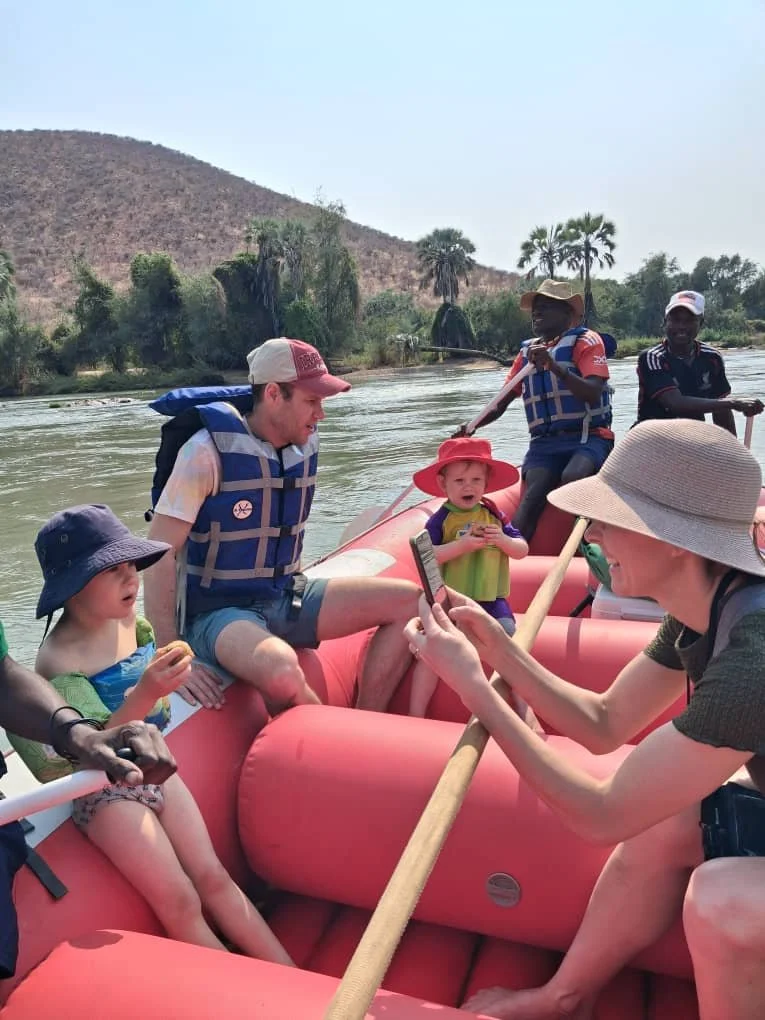 People on a whitewater rafting boat on a calm river with lush hills and palm trees in the background.